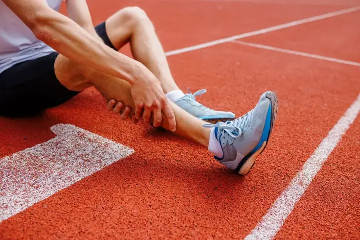 Athlete sitting on a running track, holding his lower leg in pain, possibly due to a sports injury.